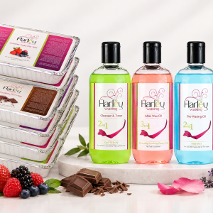 Harley Waxing product range displayed in a studio setting, featuring stacked hot wax trays and three bottles of cleanser and toner, after-wax oil, and pre-waxing oil, surrounded by fresh berries, chocolate, lavender, rose petals, greenery, and a rolled white towel.